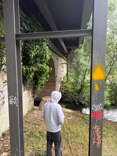 A man wearing a grey hoodie stands beside a storm drain, near a railing covered in graffiti. The railing is painted dark grey and has yellow, red, white, and orange graffiti on it. Beyond the railing, ivy grows on a stone structure. A stream runs beneath the stone structure. The image was taken in Bragança, Portugal.