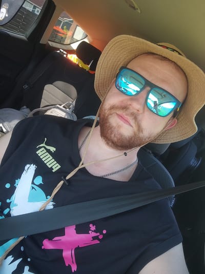 A man with a beard and a straw hat is taking a selfie inside a car. He is wearing sunglasses and a black t-shirt with a Puma logo and a colorful splatter design. A seatbelt is visible across his chest. The background shows car seats and windows with a view of houses and trees. The image is taken in Zashchita, Russia.