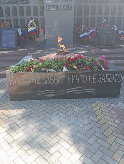 The image depicts a memorial site with an eternal flame and floral tributes. The central monument features the years "1941-1945" prominently displayed. Surrounding the monument are numerous plaques listing names, likely of fallen soldiers. Several wreaths, adorned with red, white, and blue ribbons, are placed around the memorial. In the foreground, a dark granite structure bears the inscription "НИКТО НЕ ЗАБЫТ НИЧТО НЕ ЗАБЫТО" which translates to "No one is forgotten, nothing is forgotten." This structure is also decorated with a bouquet of red and pink roses. The ground in front of the monument is paved with interlocking bricks. The scene appears to be outdoors, under daylight, with some dappled sunlight and shadows cast on the ground. The memorial is located in the city of Otradnaya, Russia. There are no people visible in the image.