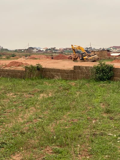 The image depicts a construction site in the city of Kwang, Nigeria, characterized by an overcast sky and a landscape dotted with unfinished buildings and mounds of red earth. In the mid-ground, a yellow excavator is parked. To the left of the excavator, two individuals are visible, one in dark clothing and the other in a yellow hard hat, standing near piles of dirt. Further in the background, a residential area with multiple houses, some appearing to be under construction and others completed, stretches across the horizon. The foreground is dominated by overgrown grass and a low brick wall.