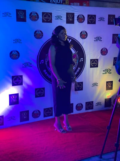 Here is a description of the image:
A woman poses for a photo in front of a backdrop decorated with logos and text, including "DOUBLE TWIN," "D-RICH," "MORALES," and "DMW WORLDWIDE." She is wearing a dark dress and heels. The lighting appears to be a mix of blue, red, and yellow. The location is in Iranla, Nigeria.