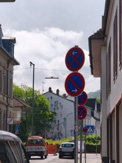 The image depicts a street scene in Lörrach, Germany, under a partly cloudy sky. In the foreground, several traffic signs are prominently displayed, including three circular signs with red borders and blue backgrounds, indicating parking restrictions with directional arrows. The topmost sign shows a blue 'X' with a left arrow, signifying no stopping in either direction. Below it, a sign with a diagonal red bar and a right arrow indicates no parking. Another similar sign is partially visible at the bottom. Adjacent to these is a blue square sign with a pedestrian crossing symbol.

On the right side of the street, a building with white walls and brown window frames is visible. On the left, older buildings with stone facades and Mansard roofs are present. In the mid-ground, an orange utility truck is parked behind a yellow construction barrier. A white car is also visible. Behind the truck, a building features a large mural depicting numerous stylized human figures walking. Trees with vibrant green foliage line the street, and power lines crisscross the sky. The street itself is paved, and a sidewalk is present on the right.