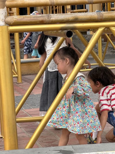 The image shows a group of children in an outdoor setting, likely a walkway or recreational area, in Hội An Ward, Vietnam. The children are interacting around a golden metal structure, possibly part of a playground or a decorative fence.

The foreground is dominated by the golden metal bars, some of which are cylindrical, and a bamboo pole. In the middle ground, a young child wearing a white top and a colorful floral dress is leaning forward, looking down. Next to this child is another child wearing a red and white striped shirt and blue shorts, also looking downwards. There is a third child partially visible in the background, wearing a blue and white patterned top and shorts.

The ground beneath them appears to be paved with bricks in a decorative pattern. The overall lighting suggests it is daytime, with no indication of specific weather conditions. The children's posture and focus suggest they are engaged in an activity together, perhaps playing a game or observing something on the ground. There is no visible text in the image. The scene conveys a sense of youthful energy and interaction.