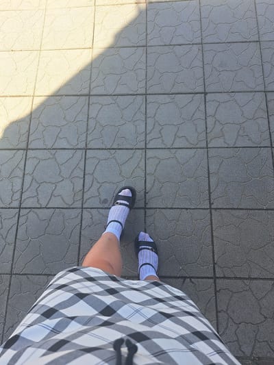 The image shows a first-person perspective of someone standing on a paved outdoor area. The ground is covered in square tiles with a textured, irregular pattern. A diagonal shadow casts across the tiles from the upper left to the lower right of the frame. The person is wearing a black and white checkered skirt, white socks with thin blue stripes, and black sandals. The scene appears to be a public walkway or sidewalk in Leningradskaya, Russia, likely on a sunny day given the distinct shadow.
