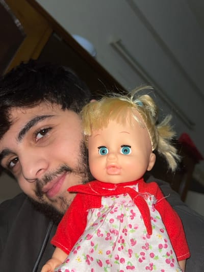 A man with dark hair and a beard holds a doll with blond hair, blue eyes, and a red and white floral dress. The man looks up at the camera with a slight smile. The doll's face is positioned beside the man's face. The background includes a glimpse of a ceiling with lights and a door. The man and the doll appear to be the main subjects, with the rest of the background out of focus.