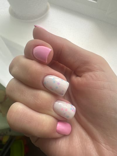 The image displays a close-up of a person's left hand, showcasing manicured nails. The nails on the thumb, index finger, and pinky finger are painted a solid pastel pink. The middle and ring fingers have a white base coat adorned with small, scattered pastel dots in pink, blue, and yellow. The hand is positioned against a translucent white or light-colored fabric, possibly a sheer curtain, with visible grid patterns. Natural light appears to be coming from the right, casting a soft illumination on the hand and creating subtle shadows. The background is out of focus and does not provide any discernible details about the location. There are no indications of the city of Sayanogorsk, Russia, or any other specific geographical information within the image.