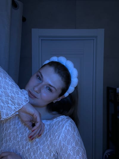 A woman with light skin and a white, puffy headband tilts her head to the side, looking at the camera. She wears a white lace top with long, flared sleeves. Her right hand, with red nail polish, is near her face. Behind her is a closed, white door with rectangular panels. The lighting is dim and blue-toned, creating a calm and intimate atmosphere. The image is taken at an unusual angle, potentially upside down.