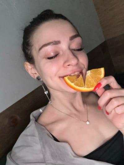 A female individual is depicted in an indoor setting within Reutov, Russia. Her face is in the upper half of the frame, with her eyes closed or partially closed and her mouth open, biting into a segmented orange. Her right hand holds the orange. She has dark hair pulled back, visible eyelashes, and shaped eyebrows. Her left earlobe features a plug or tunnel, from which a long, dangling earring with multiple clear, cube-shaped beads extends. A thin necklace with a small, clear pendant rests on her collarbone. Her fingernails are painted red. She wears a black garment underneath a light gray or muted-colored outer garment draped over her shoulders. The background is dim and indistinct, with a vertical wooden element visible to the right and a solid-colored wall to the left. The lighting is low, illuminating the subject primarily from the front.