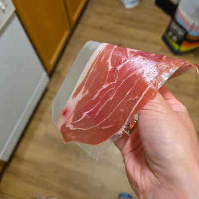 In this close-up shot, a person's hand is holding a thin slice of cured ham, possibly prosciutto, which is draped over a clear plastic separator. The ham is a deep pinkish-red with visible marbling of white fat. The hand, with fair skin and visible creases, is gently gripping the ham, suggesting it is being prepared or presented.

The background is blurred, but hints of a domestic setting are discernible. To the left, out of focus, is a colorful object that appears to be part of a packaging or label, displaying stripes of rainbow colors including pink, orange, yellow, green, and blue, against a dark background with some white text or symbols. This colorful element might be associated with the packaging of the ham or another item nearby.

The floor surface behind the hand and ham is a warm, light brown wood grain, suggesting a table or countertop. Further in the background, a sliver of a white surface is visible, potentially another countertop or piece of furniture.

The lighting appears to be ambient, casting soft shadows and highlighting the texture of the ham and the details of the hand. There are no strong indications of time of day or weather, but the indoor setting suggests it could be anytime. The focus is entirely on the ham and the hand, creating an intimate and perhaps appetizing scene, with no discernible emotions expressed. The location context is Portland, United States of America, though no specific visual cues in the image directly confirm this.