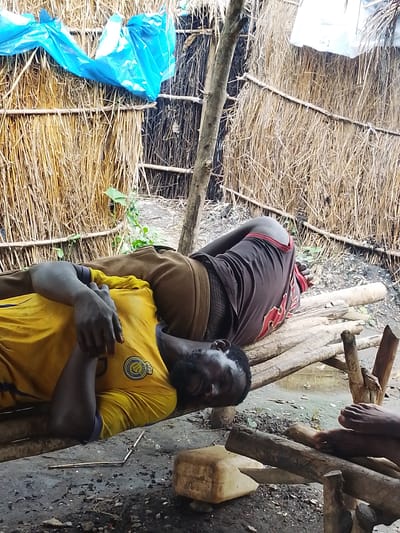 The image depicts a crude shelter constructed from dried grasses and wooden poles, suggesting a rural or impoverished setting in Otulu, Nigeria. Two individuals are resting on a makeshift platform made of wooden planks. One person, wearing a yellow t-shirt with a circular emblem and dark blue trim, is lying on their back, with their head resting on their arm and their eyes closed. Their hands are clasped over their chest. The other person, lying on their side, is wearing a brown long-sleeved shirt and dark shorts with red trim. Their body is partially obscured by the first individual. The ground beneath the platform is rough and uneven, covered with dirt, small stones, and debris. To the left of the shelter, a bright blue plastic sheet is draped over the grass wall, possibly for protection from the elements. In the background, more dried grasses and wooden supports form the structure of the shelter. The lighting appears to be natural daylight, creating soft shadows within the shelter. The overall impression is one of simple, basic living conditions.