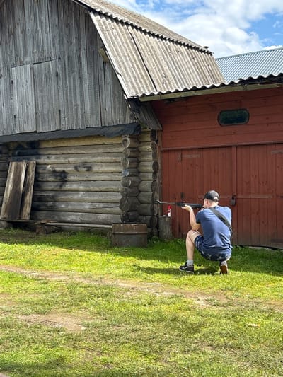 The image depicts a person crouching in front of a rustic wooden building, holding a rifle. The person is wearing a blue t-shirt, dark shorts with a pattern, a black baseball cap, and sneakers. They are aiming the rifle at something out of frame. The building appears to be made of weathered logs and has a reddish-brown wooden section with an oval window. Next to it is another section of the building with grey, corrugated roofing and wooden siding. The scene is set outdoors on a sunny day, with green grass covering the ground. The person's stance and the rifle suggest an activity related to shooting or target practice. The overall atmosphere is casual and outdoorsy, set in a rural location. The location context provided is Дресвяново, Russia, which aligns with the rustic architecture and natural setting. There is no visible text in the image.