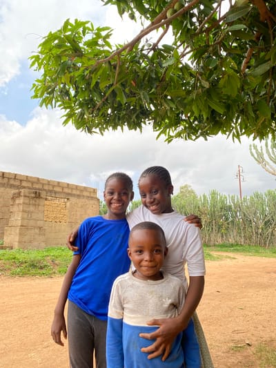 Three smiling African children pose closely together in an outdoor setting in Kuru Dungus, Nigeria. A girl with braided hair wears a white t-shirt and places her arm around the child in the center. The central child, a boy, wears a gray and white shirt. The child on the left wears a bright blue t-shirt. They appear to be standing next to a wall constructed of tan blocks. A large tree with green leaves partially obscures the left side of the frame. In the background, utility poles and tall, cactus-like plants are visible. The ground is a mix of dirt and grass. The sky is cloudy.