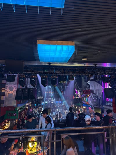 The image depicts a crowded indoor venue, likely a nightclub or entertainment establishment, located in the city of Thủ Đức, Vietnam. The atmosphere is illuminated by vibrant, colorful lights, including a large, rectangular blue lighting fixture suspended from the ceiling. The venue features a multi-level design with numerous patrons occupying various areas.
In the foreground, a metal railing separates the viewer from a group of individuals seated at tables laden with drinks and food. One person in the foreground is holding a red object. Further back, several people are standing and interacting. Notable figures include a man in a white shirt with his arm around a woman, and another man in a dark polo shirt.
The midground reveals a densely packed crowd of people standing and moving. Above them, stage lighting equipment, including spotlights and speakers, is mounted on trusses. Behind the crowd, a large, decorative structure resembling a casino slot machine with flashing neon lights is visible. This structure is adorned with playing card symbols, including spades and clubs.
The background shows more of the crowded venue, with additional lighting and what appears to be architectural elements. The overall impression is one of a lively and energetic nightlife scene.
