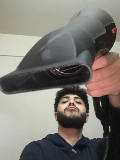 A man holds a gray hair dryer close to the camera. He has a beard and is wearing a navy blue sweatshirt. The hair dryer is angled downwards. The setting appears to be indoors with a light-colored wall as the background. The man's face is visible with a slight smirk.