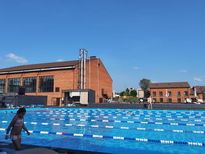 A wide outdoor swimming pool with lane markers is in the foreground, with a bright blue sky and some scattered clouds above. A young girl in a patterned swimsuit is walking on the pool deck to the left of the frame. In the background, there is a large brick building with many windows, and another smaller brick building to the right. Several people are visible in and around the pool. The location appears to be a sports or recreational complex in Pirogovo, Russia.