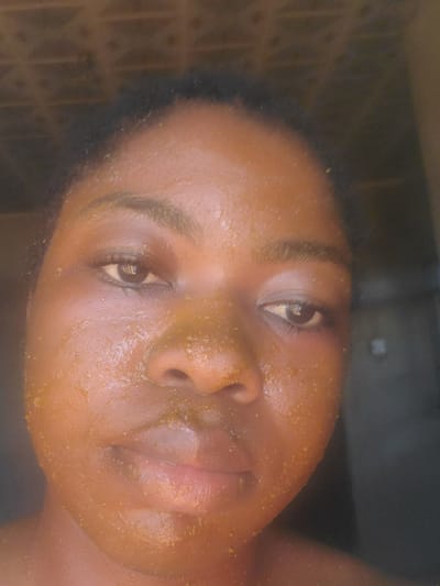 A close-up portrait of a dark-skinned individual, likely a young adult female, is depicted in Kakau, Nigeria. The subject faces the camera directly, maintaining eye contact, with a neutral expression. Their face is entirely coated in a granular, yellowish-orange substance, consistent with a facial mask or scrub, covering the forehead, cheeks, nose, and chin. Small specks of the substance are visible along the hairline and upper chest. Both eyes are open, and reflections of an unidentified light source are present in the pupils. The hair, dark and short, is visible at the top of the head. The background is dark, out-of-focus, and appears to be an interior wall or doorway with subtle, barely discernible horizontal textures. The lighting is frontal and bright, illuminating the subject against the subdued background.