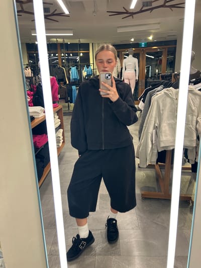 A young woman is captured in a mirror selfie, standing in what appears to be a Lululemon clothing store in Whistler, Canada. She is dressed in an all-black outfit, consisting of a zip-up hoodie, wide-leg pants, white crew socks, and black sneakers featuring a distinct white emblem on the side. She holds a smartphone with a light-colored case up to her face, eyes closed as she takes the photo.
The store interior is brightly lit by horizontal fluorescent tube lights, reflecting prominently in the mirror. The flooring is grey tile. In the background, various clothing items are displayed on wooden and metal racks, including athletic wear in shades of white, grey, black, blue, and pink. Mannequins wearing white and pink clothing are visible further back. A small white tag on a clothing item on a shelf clearly shows a partial "lululemon" logo. A green "EXIT" sign is also visible in the reflection above a doorway. To the left, a decorative wooden coat rack with multiple branches adds to the store's modern aesthetic. The scene suggests a retail environment during daytime hours, well-illuminated indoors.