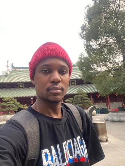 A selfie captures a young Black man outdoors in what appears to be a traditional Chinese temple complex in Guangzhou, China. He looks directly at the camera with a neutral expression. He wears a red knit beanie and a black t-shirt with "BALENCIAGA" written across the front in a stylized font with blue and red outlines. Grey backpack straps are visible on his shoulders.

The background features striking traditional Chinese architecture, including a large building with an intricate green tiled roof and red pillars. On the building's facade, vertical Chinese characters are visible, likely reading "天王殿" (Hall of Heavenly Kings), a common structure in Buddhist temples. Surrounding the architecture are lush green trees, including a dense canopy overhead and manicured bushes. Stone railings and what appears to be a traditional incense burner or offering pot are also present near the building. The lighting is diffused, suggesting it is daytime under an overcast sky.