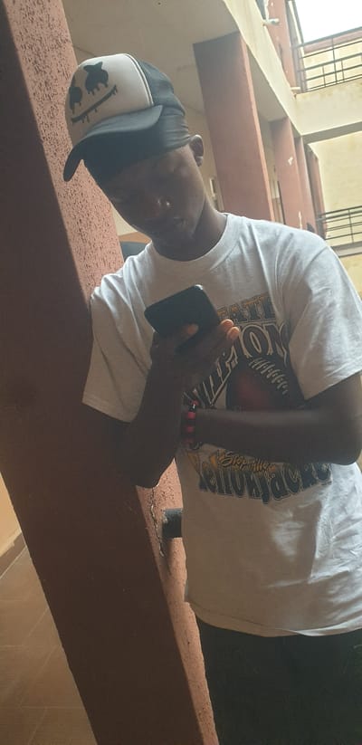 A young person wearing a white t-shirt with a graphic print and a black and white trucker hat with a melting smiley face logo is leaning against a brown textured pillar. The person is looking down at a black smartphone in their hands. They are wearing a black durag under the hat. The background shows a building with a pale yellow facade and dark metal railings on balconies. The flooring visible in the lower left is tiled in a light brown color. This scene appears to be an outdoor common area or hallway within a building in Yelwa, Nigeria.