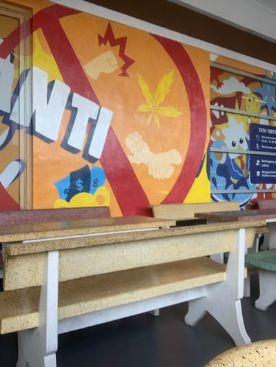 The image shows a public space with a mural on a wall depicting a stylized red circle with a line through it. The large stylized letters "ANTI" are painted in white in the circle. The mural has a background of orange and yellow with cartoon depictions of a fist, a marijuana leaf, and two hands shaking. The scene includes two concrete benches and tables. The benches are made of rough concrete with a tan top and a white base. The tables are made of rough concrete with a red top and tan bottom. The scene is outdoors in the city of Malang, Indonesia (coordinates: -7.938, 112.626). The location is a public plaza or park.