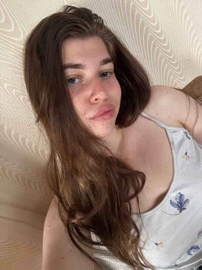 The media file shows a close-up, overhead shot of a young woman lying on her back. She is wearing a white tank top with a subtle floral pattern in blue. Her long, brown hair is spread out around her head and shoulders. The background appears to be a textured, light-colored surface, possibly a bed or sofa, with a repeating wave-like pattern. The lighting suggests it is daytime, possibly indoors with natural light. The woman's face is partially visible, with her eyes open and looking towards the camera. Her expression is neutral. There is no visible text in the image. The location context provided is Мещёры, Russia.
