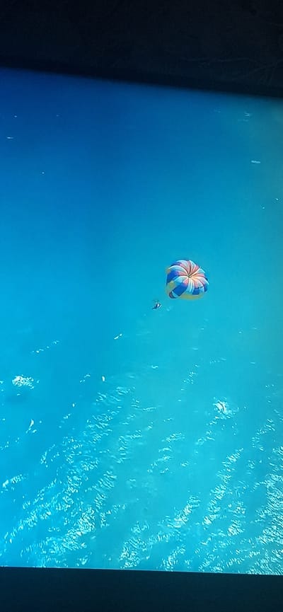 The image shows a brightly colored parachute, likely used for parasailing, floating in a clear blue sky over a body of water. Two figures are visible beneath the parachute, appearing to be participants in the activity. The water below has a rippled texture, suggesting a calm surface with some light waves. The image is taken from a high vantage point, looking down at the scene. There is no indication of the city of Bila Tserkva, Ukraine, or any urban elements within the frame. The top portion of the image is dark, possibly indicating the edge of a screen or a dark background.
