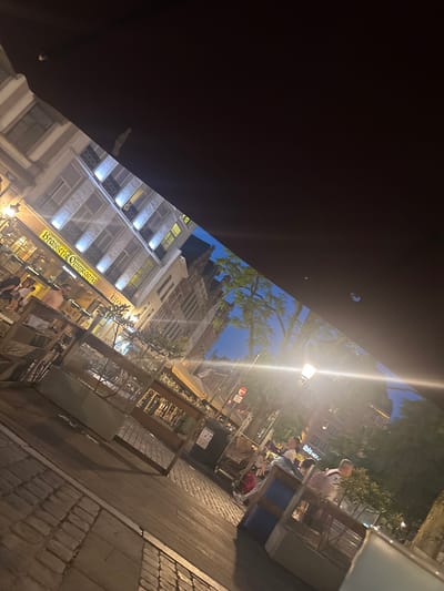 An exterior shot shows a street scene at night in Brussels, Belgium. A prominent awning covers part of the scene, casting a shadow. In the background, buildings line the street, with lights illuminating windows. One building features signage that reads "Brasserie Ommegang." Other signs in the background are partially visible, including "ibis HOTEL." Several people are seated at outdoor tables, with some appearing to be dining or socializing. A streetlamp with a warm glow illuminates the area. The street is paved with cobblestones, and some trees with green foliage are visible. A trash receptacle is situated on the sidewalk.