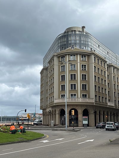 This image captures an urban street scene in Gijón/Xixón, Spain, under an overcast sky. The prominent subject is a large, multi-story building dominating the left side of the frame. Its architecture features a beige lower section with multiple rows of rectangular windows and a ground floor with a series of arched openings. The building's upper section transitions into a contemporary curved glass facade, reflecting the cloudy conditions.

On the right, a street runs parallel to the building, featuring clear white arrow markings on the asphalt indicating traffic direction. A central landscaped median strip separates the roadway. On this median, two individuals are engaged in gardening. They are dressed in high-visibility work attire, one in green and the other in orange, and appear to be tending to plants and flowers in the grassy area.

Several cars are visible on the street, including a white van with a license plate partially visible as "KXX" and a dark car with a license plate partially visible as "LYY". Traffic lights displaying yellow/amber are present, as is a large information board. Text visible on this board reads "Concejo de Gijón" (Council of Gijón), along with what appears to be a map or descriptive text about the locale. The sky is uniformly grey and cloudy, indicating an overcast daytime.