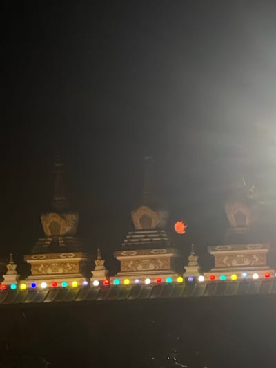The image depicts a nighttime scene in Pokhara, Nepal, featuring the reflection of illuminated structures in water. The structures appear to be temples or monuments, characterized by ornate designs and vertical rows of colored lights (red, blue, yellow, white, and green). An orange glow, possibly from the moon or a distant light source, is visible in the background. The overall atmosphere is dark and slightly hazy, with the water surface reflecting the lights and structures, creating a mirrored effect. The sky is dark. The image is oriented such that the reflection appears upright and the actual structures are inverted.