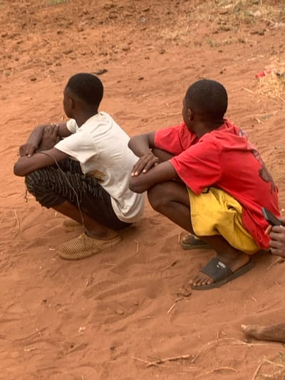 Two boys are crouched on dirt ground. The boy on top wears a red t-shirt and yellow shorts, while the boy below has a white t-shirt and dark patterned shorts. Both have their heads bowed and arms wrapped around their legs. The feet of the top boy wear dark sandals. The bottom boy wears light colored, closed-toe sandals. The dirt ground is covered in scattered dry grasses.