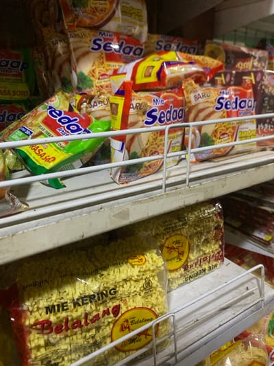 The image shows the interior of a refrigerator, stocked with various packaged instant noodles. The refrigerator is white with rusty metal shelves. On the left side, there are multiple packages of "Mie Sedap" brand instant noodles with different flavors, all featuring a red and yellow color scheme and images of noodles, chicken, and vegetables. On the right side, there are two packages of "Mie Belalang" brand instant noodles with a yellow and red color scheme and images of noodles and a logo. The noodles in the packages appear to be dry, curly, and yellow. The packages are arranged vertically and horizontally on the shelves, with some partially obscured by others. The image was taken in a store in Kecamatan Lawang, Indonesia, located at latitude -7.829 and longitude 112.696.