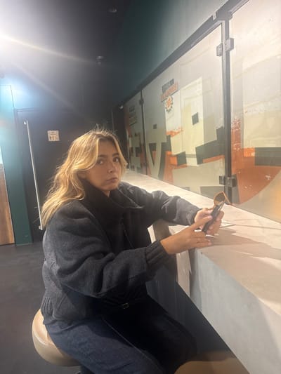 An interior image depicts a female individual seated at a counter, likely within the lobby or snack area of a UGC Ciné Cité cinema, specifically UGC Ciné Cité Carré de Soie in Vaulx-en-Velin, France, as indicated by the "UGC FLAMMES" logo on the background wall. The subject, a woman with shoulder-length blonde hair, appears to be in her 20s or 30s and is wearing a dark gray, high-collared jacket or sweater, dark blue jeans, and a dark chain-strap shoulder bag. She is positioned on a round, brown stool, facing the camera with a neutral expression. Her left hand holds a dark smartphone, while her right hand holds a pair of brown-framed sunglasses, both resting on a light gray, possibly concrete, counter. The background features a wall composed of large panels adorned with a muted orange, beige, and black geometric graphic design. To the left, a dark door with a white restroom symbol is visible. The ceiling is dark, with recessed lighting, and a prominent lens flare is present in the upper left quadrant of the image.