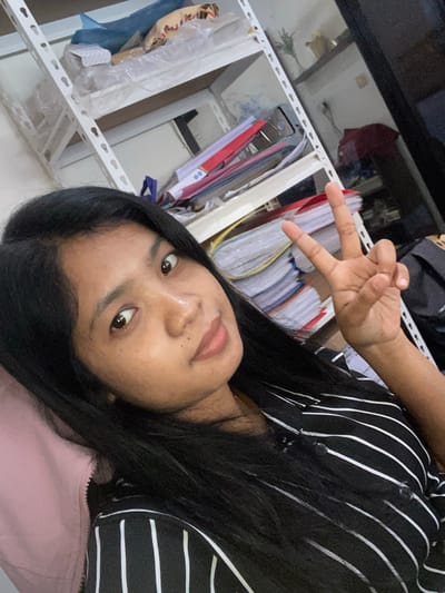 A person with dark hair is shown from the chest up, wearing a black and white striped shirt over a light pink garment. They are making a "peace" or "victory" sign with their right hand. Behind the person is a white metal shelving unit. The shelving unit is partially filled with file folders and objects wrapped in plastic. A dark reflective surface, possibly glass or polished stone, is visible above the shelving unit. It is unknown if this picture was taken in Negara, Indonesia.