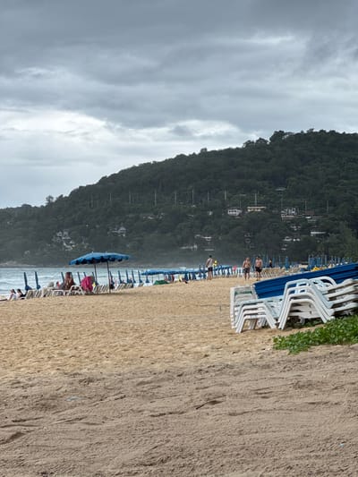 The image depicts a tropical beach scene with a lush, forested hill in the background. The foreground is dominated by a sandy beach with rows of white lounge chairs and blue umbrellas, some of which are open. Several people are visible on the beach, scattered among the chairs, some appearing to be resting or walking. The sea is visible in the distance, with gentle waves and a few people wading in the water.

The sky is overcast with soft, gray clouds, suggesting a mild or potentially rainy day. The time of day is difficult to ascertain precisely due to the cloud cover, but it appears to be daytime. The location is identifiable as Karon, Thailand, with the distinctive hillside architecture and dense vegetation characteristic of the area.

In the middle ground, a large blue umbrella is prominently placed on the beach, drawing the eye. There are also glimpses of buildings nestled into the hillside, suggesting a resort or residential area. The overall atmosphere is relaxed and tranquil, typical of a beach vacation setting, despite the cloudy weather. No text is visible in the image.