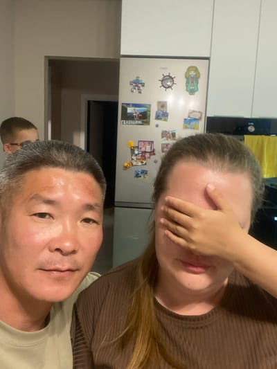 This is a close-up, selfie-style photograph taken indoors, likely in a kitchen. In the foreground, on the left, is a man with short gray hair and a light-colored t-shirt, looking towards the camera. On the right, a woman is partially visible, covering her eyes with her hand and wearing a brown ribbed shirt. In the background, a refrigerator is adorned with various magnets and pictures. Behind the man, another person, a young boy, is visible looking over his shoulder. The environment suggests a domestic setting, possibly in a residential area of Novorossiysk, Russia.
