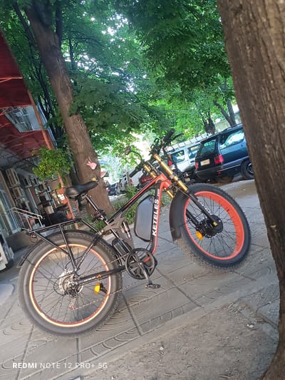 An electric bicycle is parked on a sidewalk next to a tree. The bicycle is red and black with fat tires and orange rims. It has a battery pack mounted on the frame and a rear rack. In the background, there is a street with parked cars and buildings. The image was taken in the city of Targovishte, Bulgaria.