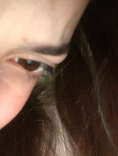 The image presents a close-up, tilted view of a person's face and hair. A portion of the forehead, eye, and nose are visible on the left side. The eye has a brown iris and noticeable eyelashes. The rest of the frame is filled with dark brown hair, with a few strands appearing lighter and reflecting light. The hair appears to be textured and layered. The angle suggests the photo was taken from a low vantage point looking upwards.
