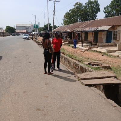 Two women are standing on the edge of a paved road in Kufang, Nigeria. The woman on the left is wearing black pants, a black tank top, and sandals. She has a black purse hanging from her right shoulder. The woman on the right is wearing a red shirt and black pants. Both women are looking at each other. The pavement is grey and appears to be freshly paved. The road is bordered by a stone retaining wall and a grassy area. There is a small wooden plank bridging a gap in the pavement. There is a line of buildings with corrugated metal roofs in the background to the right. The sky is clear and blue. The coordinates for this location are latitude: 9.863, longitude: 8.867.