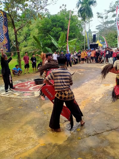 An outdoor cultural performance is captured in Tamansari, Indonesia, taking place on a wet, paved surface with puddles of yellowish water and scattered yellow leaves. A large circular symbol, painted red and white with stylized red characters, is visible on the ground.
In the foreground, a performer with their back to the viewer wears a horizontal striped shirt (tan and dark), dark pants, and a patterned kilt (dark and tan diamond design). This individual is barefoot, standing in a puddle, and holding or wearing a red, stylized horse effigy with black hair details; the word "RAHAYU" is visible in white on the effigy. Another performer, partially obscured and facing the foreground individual, is also barefoot, wearing dark pants and a shaggy brown costume element, appearing to interact with the horse effigy. To the left, a third performer dressed in black holds a long, thin pole horizontally above their head with both hands. A black bucket rests on the painted ground symbol in the mid-ground.
A crowd of approximately 30-40 spectators, including adults and youth in casual clothing, stands in a semi-circle in the background, observing the performance. The environment features dense green foliage, including banana plants and palm trees, under a bright, overcast sky. Event decorations include tall red and white vertical flags (pennants) and yellow and blue flags. Speaker stacks and additional event equipment are visible in the far right background. A black and white banner with text, partially reading "DAUN" and "ATEN," hangs from a tree on the left.