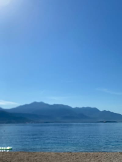 The image depicts a serene coastal scene. In the foreground, a sandy beach is visible. Beyond the beach lies a calm body of blue water, likely the Adriatic Sea. In the distance, a range of hazy, dark blue mountains forms the horizon. The sky above is a clear, bright blue with a subtle sun glare in the upper left corner. A small, partially submerged inflatable object with green and white markings is partially visible in the lower left corner of the water. The scene is tranquil and appears to be set in the city of Baošići, Montenegro, a coastal town known for its beautiful natural scenery.