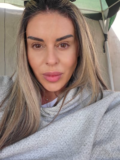 A woman with blonde and brown hair is looking directly at the camera. She is wearing a light gray hooded sweater. Her makeup includes dark eyebrows and pink lipstick. Her hair is long and appears to be windblown. The background is blurred, but a green and white patterned umbrella is visible behind her. The scene appears to be outdoors, possibly on a sunny day. There is no discernible location in the city of Ravnogor, Bulgaria.