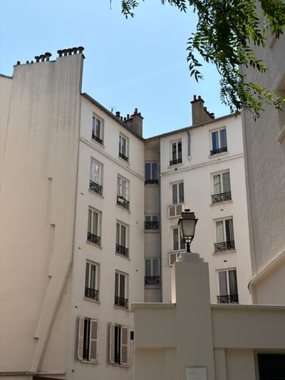 This is a low-angle, wide shot of the exterior of a white, multi-story building with a clear blue sky in the background. The building appears to be in an urban setting, possibly residential, with several windows visible on its facade. Some windows have decorative wrought iron grilles, while others have shutters. A street lamp is mounted on the side of the building. Trees with green leaves are visible in the upper left corner, partially obscuring the sky and the building. The perspective of the image is tilted, giving a sense of looking upwards. The lighting suggests it is daytime, with bright sunlight casting shadows. The image is taken from the ground or a lower vantage point, looking up towards the building. There is no visible activity or people in the frame. A small plaque is visible on the building's facade on the right side of the image, but the text is too small to read. The scene is calm and still, with no discernible emotions.