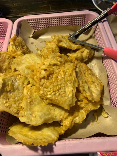 In Tanjungsepreh, Indonesia, a close-up shot focuses on a pink plastic basket filled with fried tempeh, a popular Indonesian dish. The tempeh pieces are golden brown and appear to be coated in a crispy batter. A pair of pink-handled kitchen tongs rests on top of the tempeh, with one tong gripping a piece. The basket is lined with brown paper, and the background suggests an outdoor market or food stall setting with a wooden surface visible at the top. The lighting is dim, creating a slightly shadowed effect that highlights the texture of the fried tempeh.