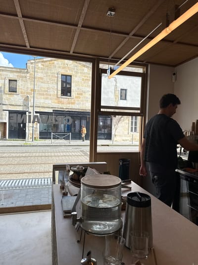 The image depicts the interior of a modern establishment, likely a café, looking out onto a street scene in Bordeaux, France.

**Interior:**
The foreground features a light-colored counter with a display of items. On the counter, from left to right, are: a small wooden stand holding a bowl of white spherical items, a larger dish with a light-colored fruit, a clear glass beverage dispenser with a wooden lid containing water and lemon/lime slices, a silver thermal carafe, and several small, clear drinking glasses. A male individual with short dark hair, wearing a dark textured short-sleeved shirt and dark pants, stands behind the counter on the right. He is positioned facing away from the camera, engaged with equipment (possibly a coffee machine) to his right, with his right hand near the controls. The ceiling is paneled with a light-colored, textured material in a grid pattern. A linear light fixture with both a warm-toned and a cooler-toned tube hangs from the ceiling. A white security camera is mounted on the upper right wall.

**Exterior (viewed through large windows):**
The street directly across features tram tracks running horizontally and is paved with cobblestones. A white metal pedestrian barrier is visible on the sidewalk. Across the street, a building constructed of light-colored, rough-hewn stone blocks dominates the view. Its ground floor houses a dark storefront with glass windows, displaying the mirrored word "GRAVAINE," indicating the establishment "GRAVAINE" at 6 Rue du Passage, Bordeaux. Several dark-framed windows are visible on both the ground and upper floors of this building. To its right, a second lighter-colored building with visible cracks in its facade is partially seen, also featuring dark-framed windows and a small, bare tree in front. One person, dressed in a light brown jacket and light pants, is visible on the sidewalk across the street, walking towards the right. A bright blue sky with some clouds is visible above the buildings.