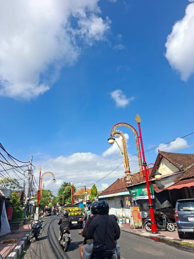 The image shows a street scene in Gianyar, Indonesia, on a bright, sunny day with a blue sky dotted with white clouds. The street is paved and has a slight incline. On the right side of the street, there are buildings with traditional tiled roofs and awnings. One building has a green banner that reads "Nonna's Cake & Bakery". There are several motorcycles and a truck on the road. The truck is carrying what appears to be bundles of yellow produce. In the foreground, two people are riding on a motorcycle, both wearing helmets. On the left side of the street, there is a sidewalk with a few buildings and some greenery. A ginger cat is lying on the sidewalk. Decorative lampposts with red poles and ornate gold accents are visible along the street. The scene depicts a typical day in a busy urban area with local commerce and traffic.