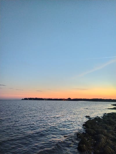 The image depicts a shoreline scene at dusk or dawn. On the left side of the frame is a light blue sky transitioning into an orange and peach-colored horizon, suggesting either sunrise or sunset. A faint contrail cuts across the sky. A dark treeline separates the sky from the water, which occupies the right side of the image. The water surface is textured with ripples and small waves, reflecting the colors of the sky. On the far right, dark, rocky outcroppings are visible along the shoreline. There are no people visible. No information supports that this scene is from Badem, India.