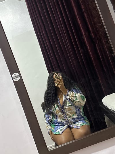 A mirror reflects a person with dark curly hair, wearing a patterned blouse, holding a phone with a zebra-striped case. The reflection also includes a portion of what appears to be a dark-colored Venetian blind and the edge of a white object. The mirror itself has a dark frame. A small round sticker, possibly a brand logo, is visible on the mirror's surface in the lower center. The location is in a room in Port Harcourt, Nigeria.