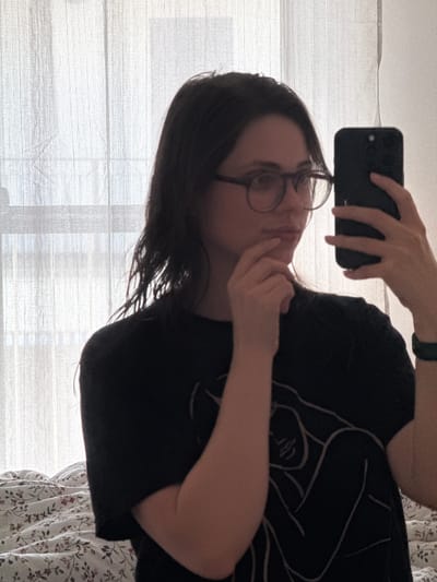 A person is holding a smartphone and taking a selfie in front of a window with white blinds. The person has dark hair, wears glasses, and a black t-shirt with a line drawing. The background also includes a bed with a floral-patterned bedspread. It is not possible to determine if the photo was taken in Balatonlelle, Hungary, as the location is not identifiable from the image.