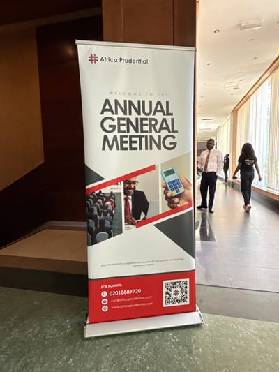 Here is a description of the image:

The image shows a banner for the Africa Prudential Annual General Meeting. The banner is primarily white with red and black accents. It features the Africa Prudential logo and contact information, including a phone number, email address, and website. There are also images relating to accounting and finance, and images of seated shareholders. The banner is standing in what appears to be an interior hallway or foyer. In the background, there are two people walking. The location appears to be Nigeria, as per the registration and regulation information on the banner. The city is not specified, and there is no indication of a city called Itirin.