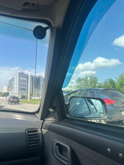 The image is a view from inside a car, looking out the passenger window. The car appears to be stopped or moving slowly in traffic on a city street. The sky is blue with some white clouds.
In the foreground, the interior of the car is visible, including part of the dashboard and door panel. A black suction cup mount is attached to the windshield, holding a thin wire. A blue wind deflector is attached to the top of the window frame.
Outside the car, a multi-story grey building with windows and signage is visible on the left side of the street. A dark-colored car is in front of the building. A dark grey hatchback is visible in the side mirror. Trees line the right side of the road.
The location is identified as Magnitogorsk, Russia, based on the provided context. The scene depicts a typical urban environment on a sunny day.